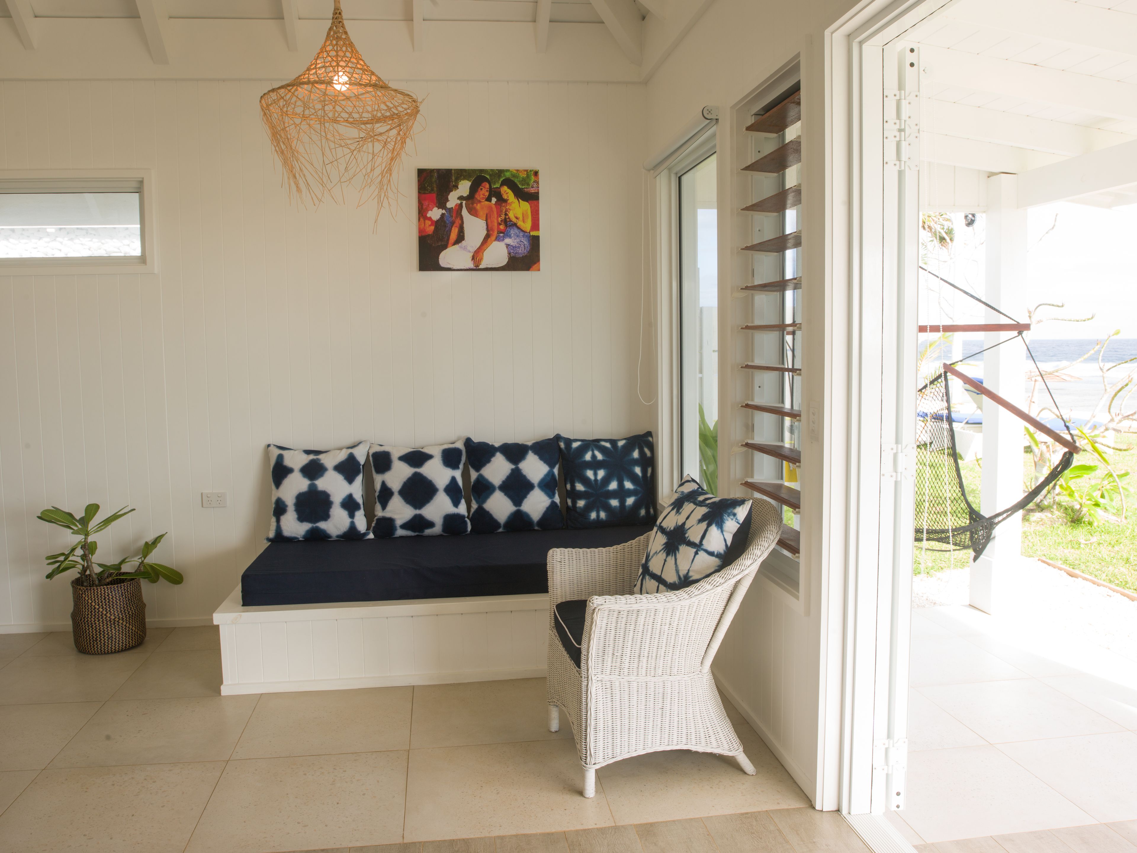 A room with a white wicker sofa, a wicker chair, a hanging light fixture, and a painting on the wall.