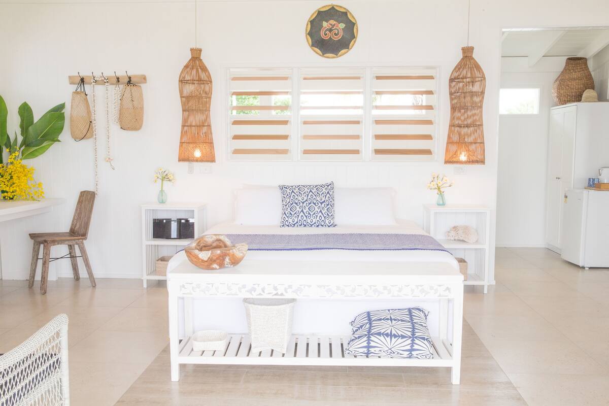 A bright, well-lit bedroom with a white bed, a wooden chair, and a small table with a basket. There are wicker lamps and a decorative wall hanging above the bed.