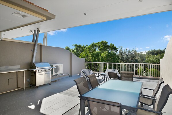 An outdoor patio with a grill, dining table, and chairs, overlooking a balcony with a view of trees. - Beachcomber on the Spit (Mooloolaba)