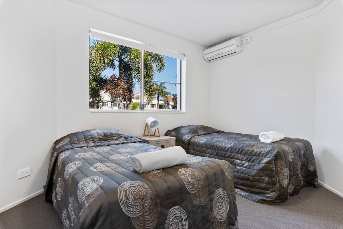 A bedroom with two beds, a window with a view of palm trees and buildings, and an air conditioning unit on the wall.