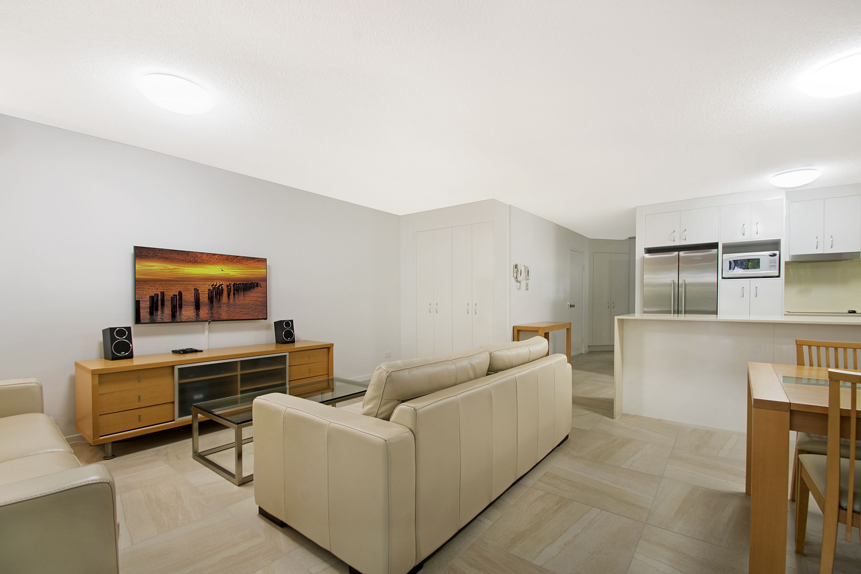 A modern living room with a sofa, a coffee table, and a TV. There is a kitchen area with cabinets and a refrigerator.