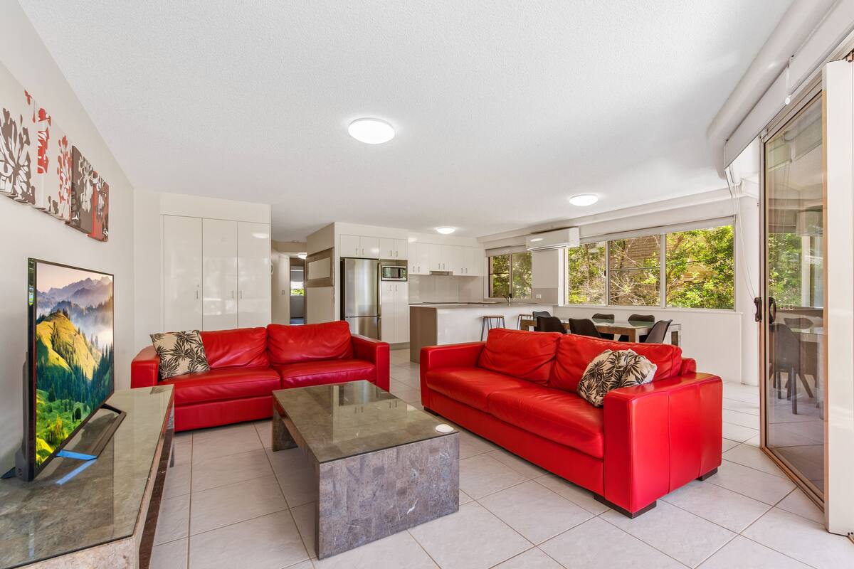 A modern living room with two red sofas, a glass coffee table, and a kitchen in the background.
