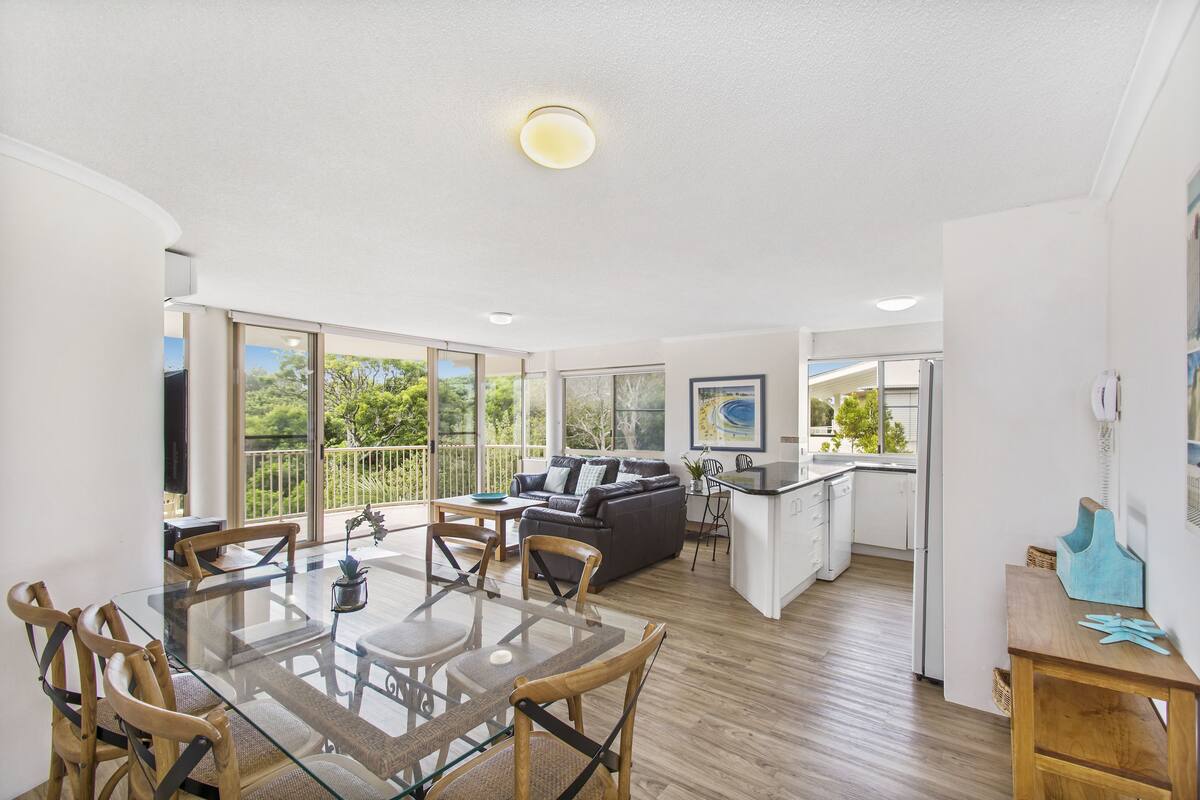 A spacious living area with a glass dining table, wooden chairs, a grey sofa, and a kitchen area with a white countertop and cabinets.