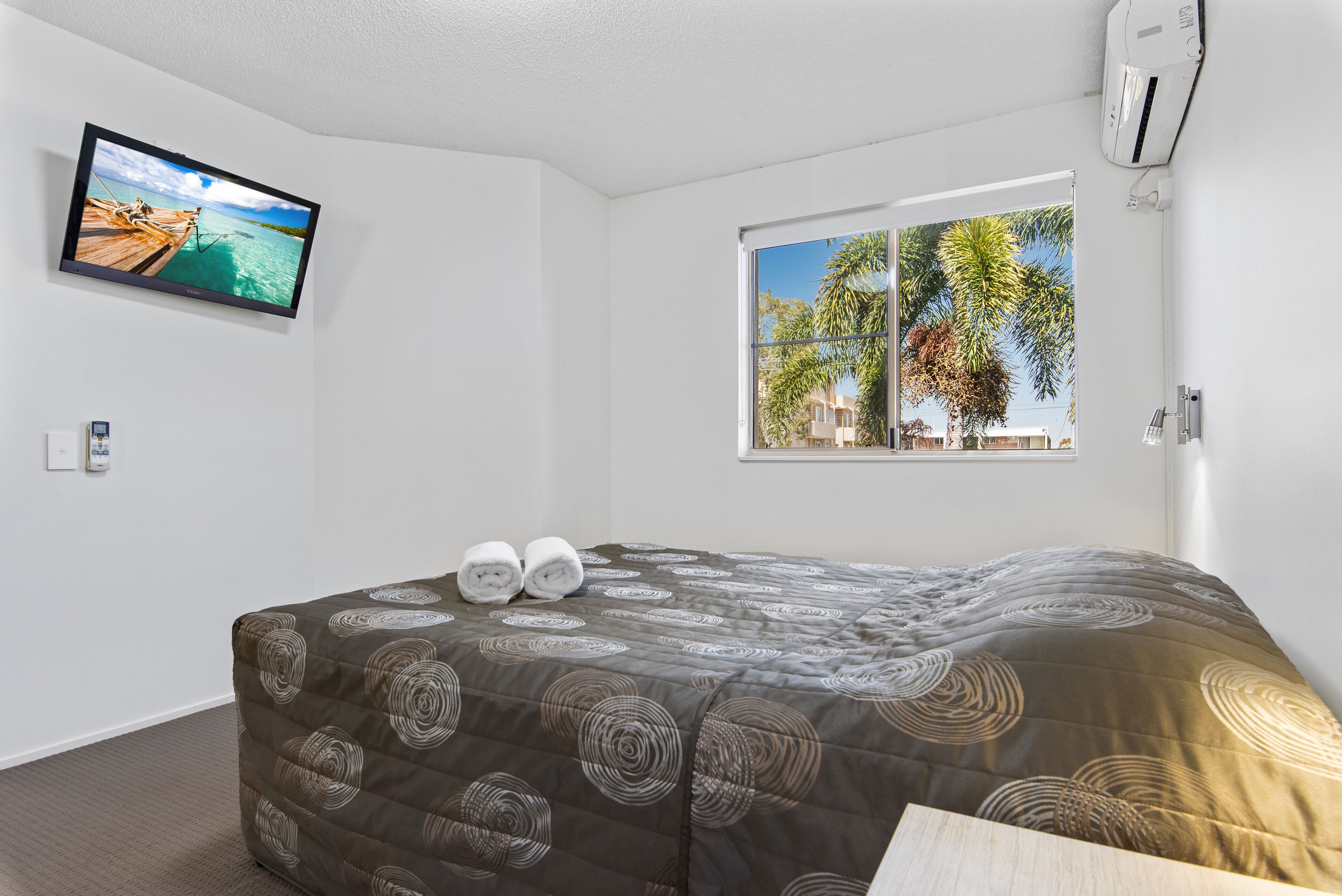 A hotel room with a bed, a TV on the wall, and a window with a view of palm trees.