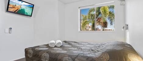 A hotel room with a bed, a TV on the wall, and a window with a view of palm trees.