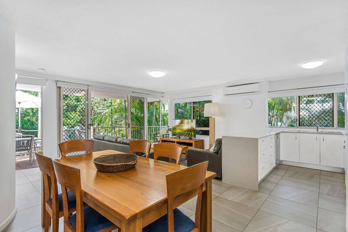 A modern kitchen with white cabinets, a dining area with a wooden table and chairs, and a TV in the living area.