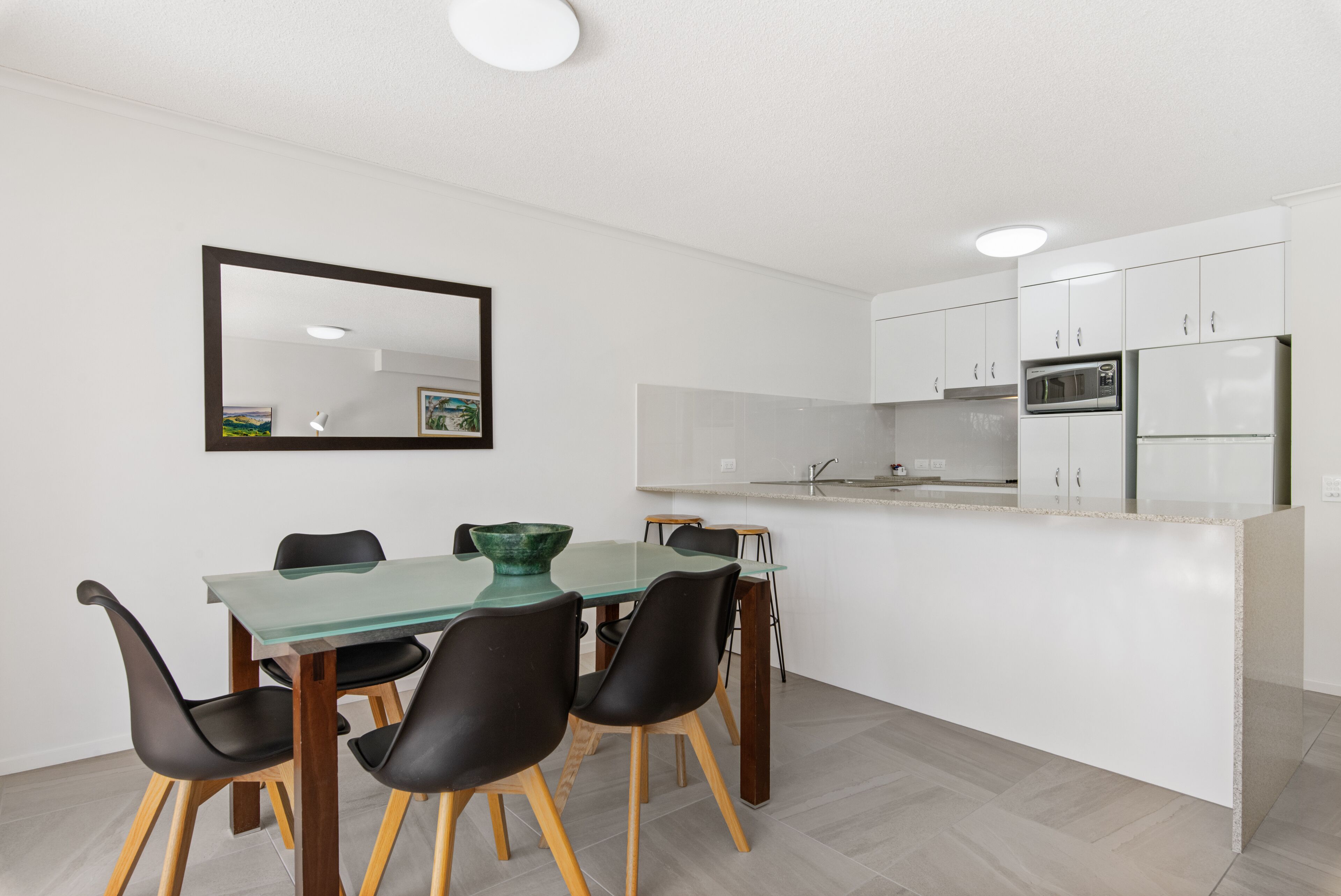 A modern kitchen with white cabinets, a microwave, and a refrigerator. There is a dining area with a glass table and black chairs.