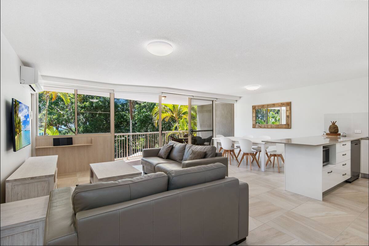 A modern living room with a grey sofa, a dining area with white chairs, a kitchen with a white countertop, and large windows with a view of greenery.