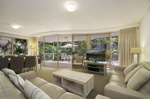 A living room with a sofa, coffee table, TV, and outdoor patio area with chairs and a table. - Beachcomber on the Spit (Mooloolaba)