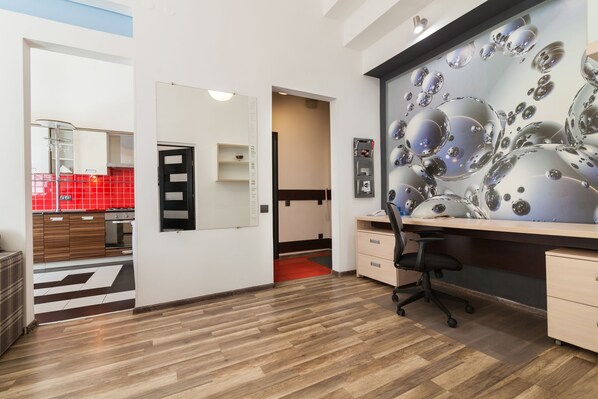 A modern office space with a wooden floor, a desk with a chair, a large mirror, and a red-tiled kitchen visible through an open door.