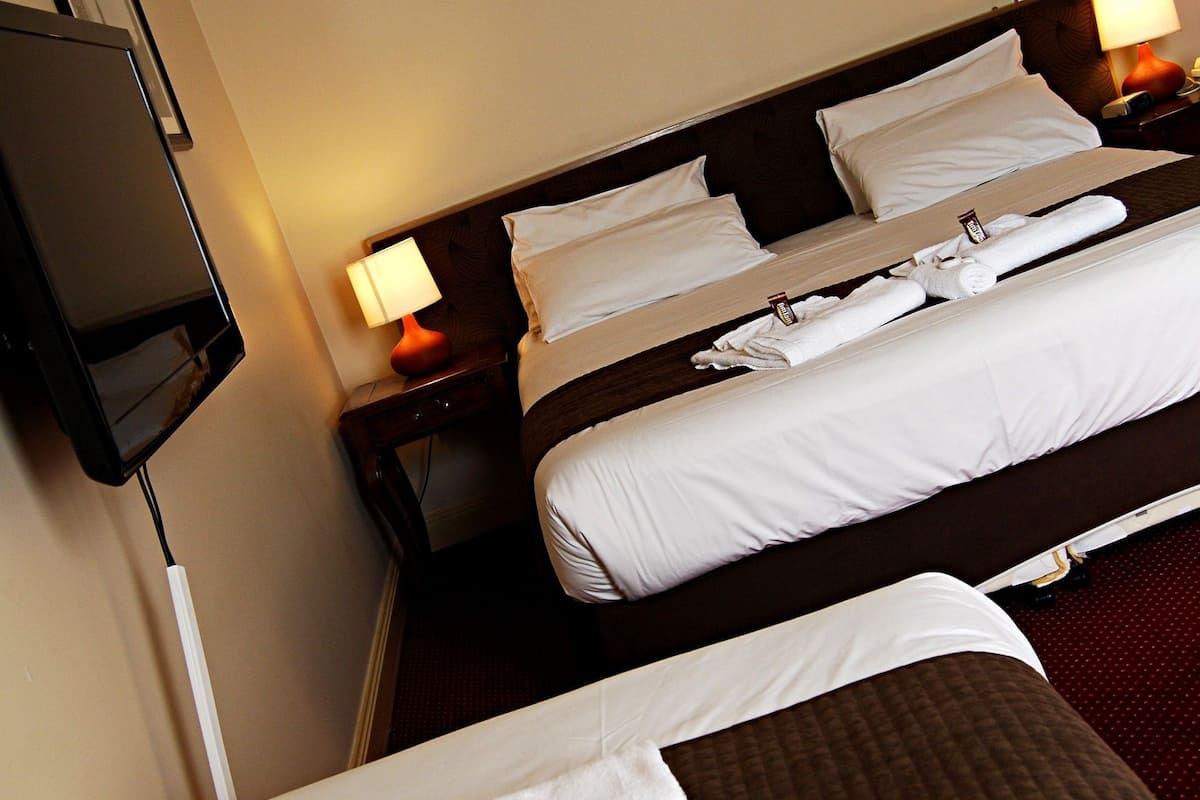 A hotel room with two beds, each with white linens and a brown headboard. There is a flat-screen TV mounted on the wall and a nightstand with a lamp.