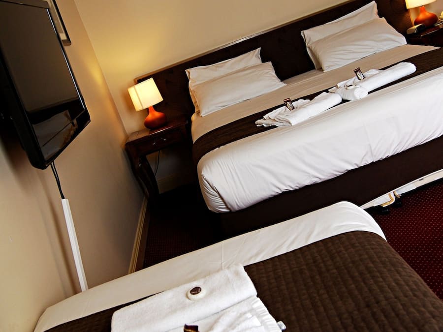 Habitación de hotel con dos camas, cada una con ropa de cama blanca y cabeceros marrones. Hay un televisor de pantalla plana montado en la pared y una mesita de noche con una lámpara.