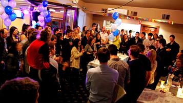 A crowded indoor event with people standing and conversing, balloons, and tables with items on them.