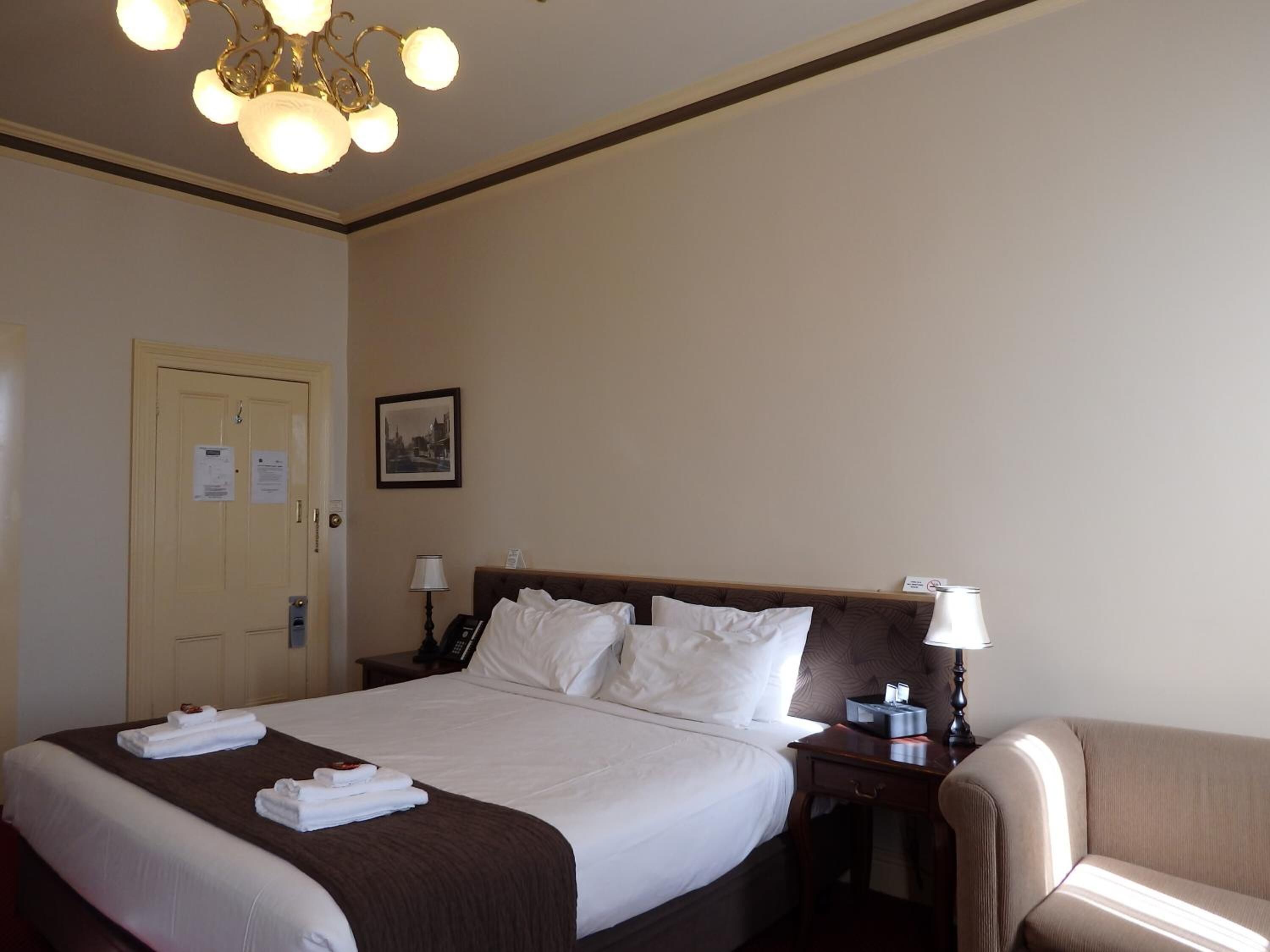 Una habitación de hotel con una cama grande, mesitas de noche, una silla y un candelabro.