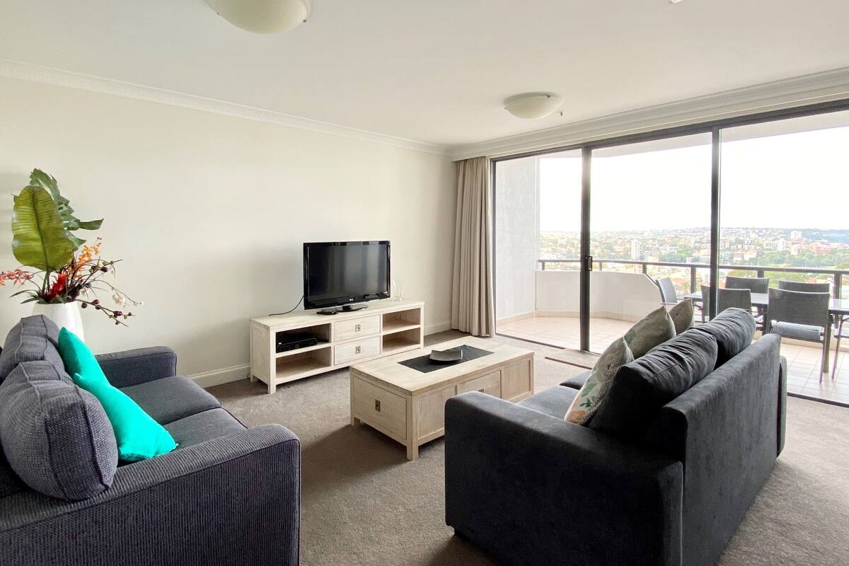 A modern living room with a flat-screen TV, two sofas, a coffee table, and a large window with a city view.