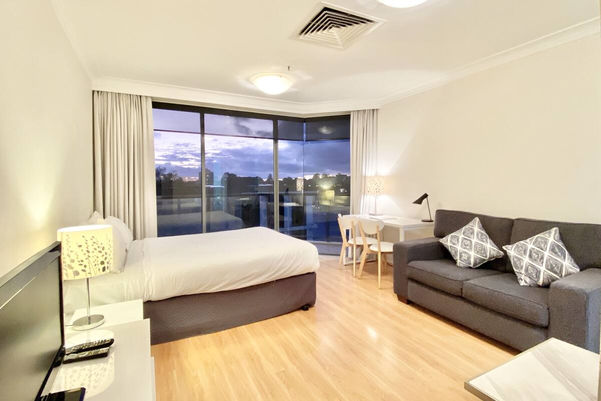 A modern hotel room with a large bed, a sofa, a desk, and a view of the city at dusk.