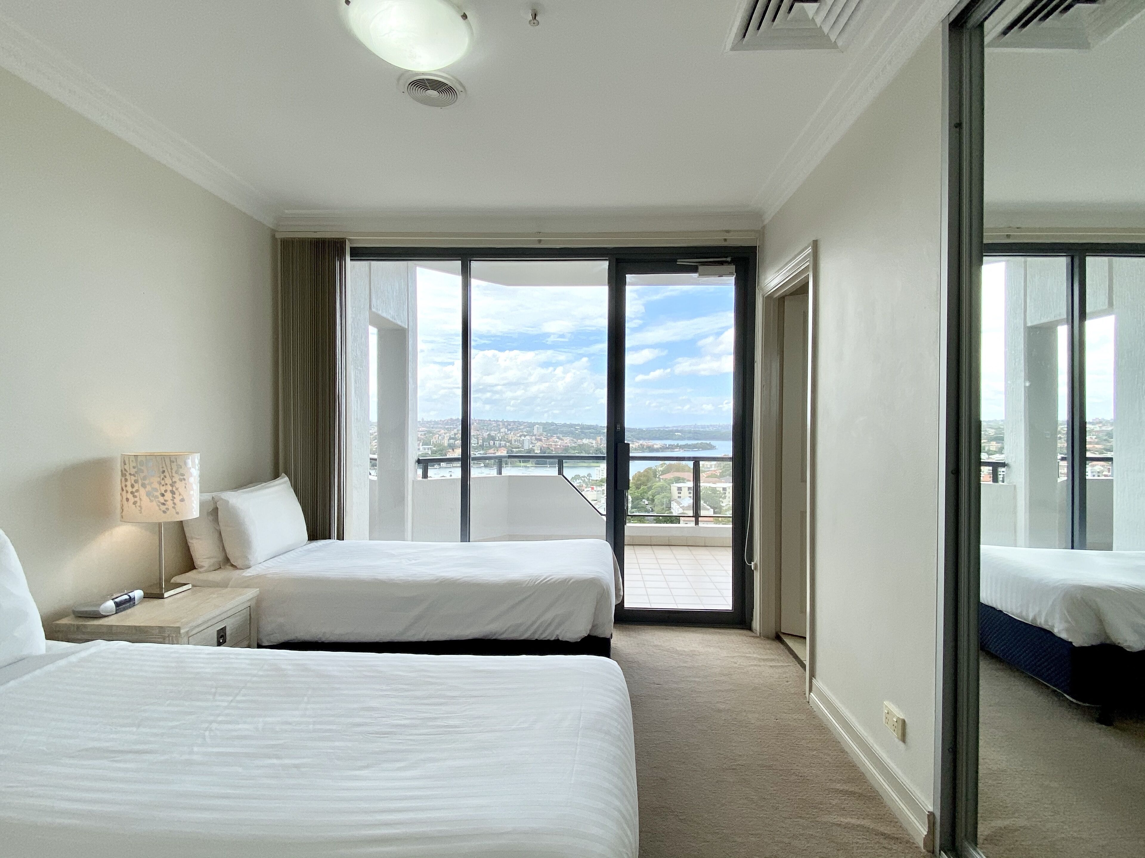 A hotel room with two beds, a bedside lamp, a view of the city through large windows, and a balcony.