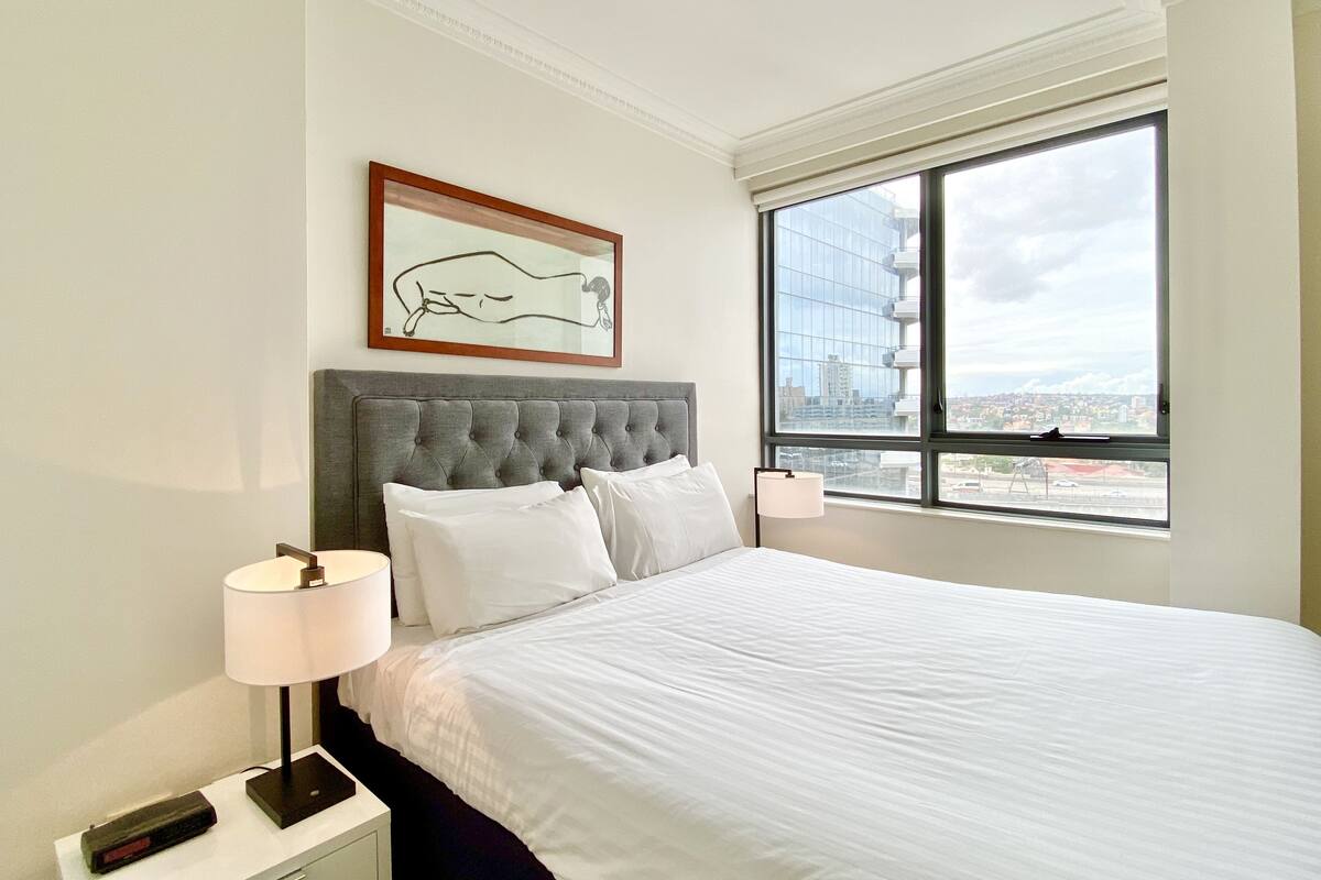 A hotel room with a large bed, a grey tufted headboard, a framed artwork, a bedside lamp, and a view of the city through a large window.