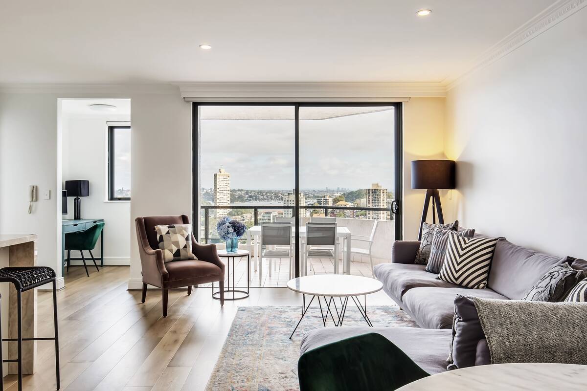 A modern living room with a sofa, armchair, and a small table. There is a balcony with a view of the city.