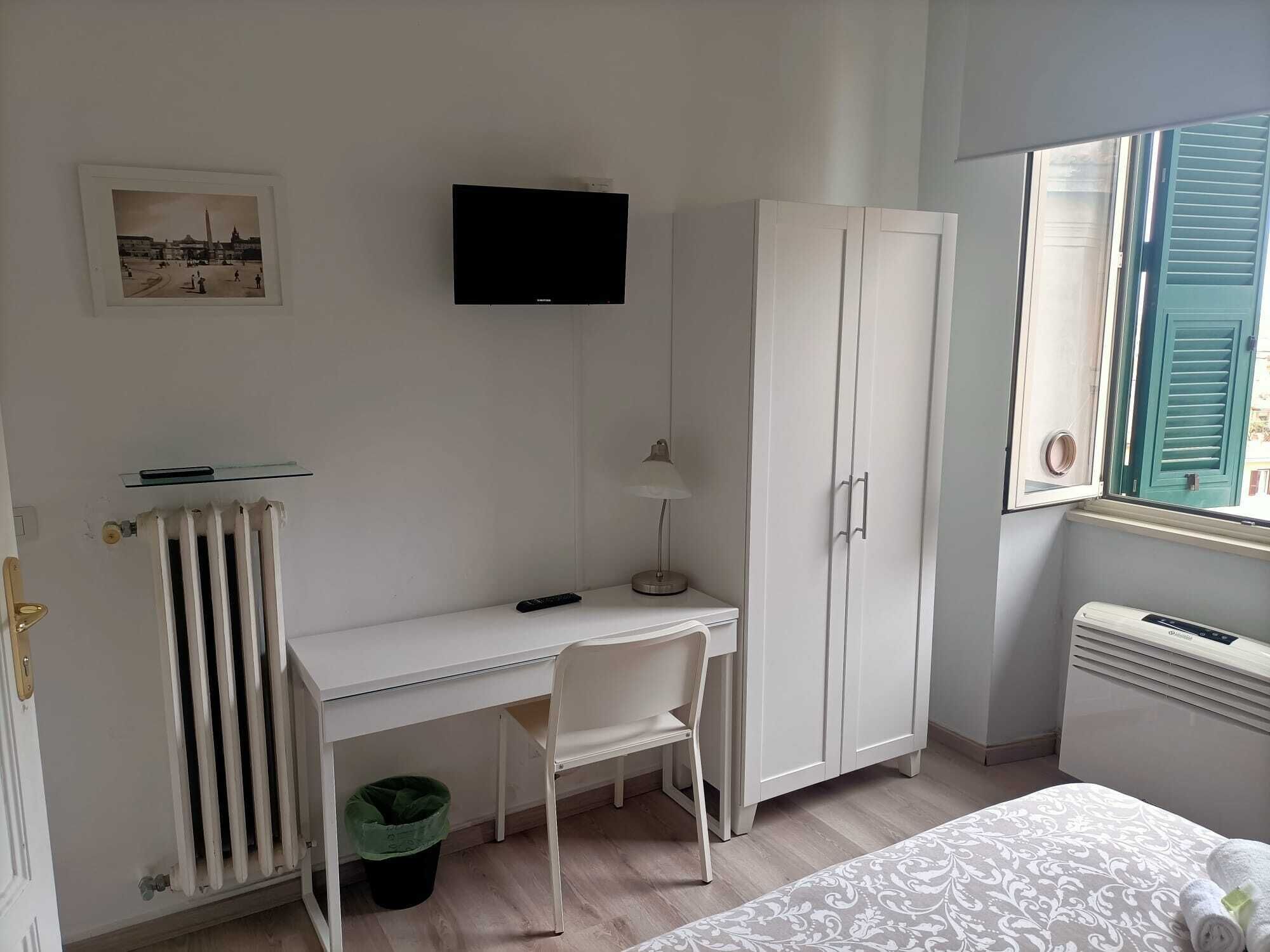 A room with a white desk, a chair, a television mounted on the wall, a wardrobe, a radiator, a window with shutters, and a bed with a patterned bedspread.
