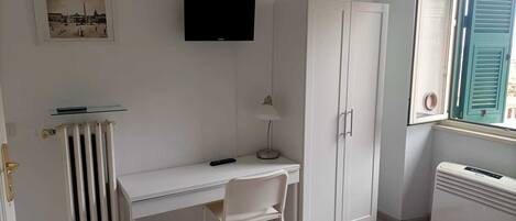 A room with a white desk, a chair, a television mounted on the wall, a wardrobe, a radiator, a window with shutters, and a bed with a patterned bedspread.
