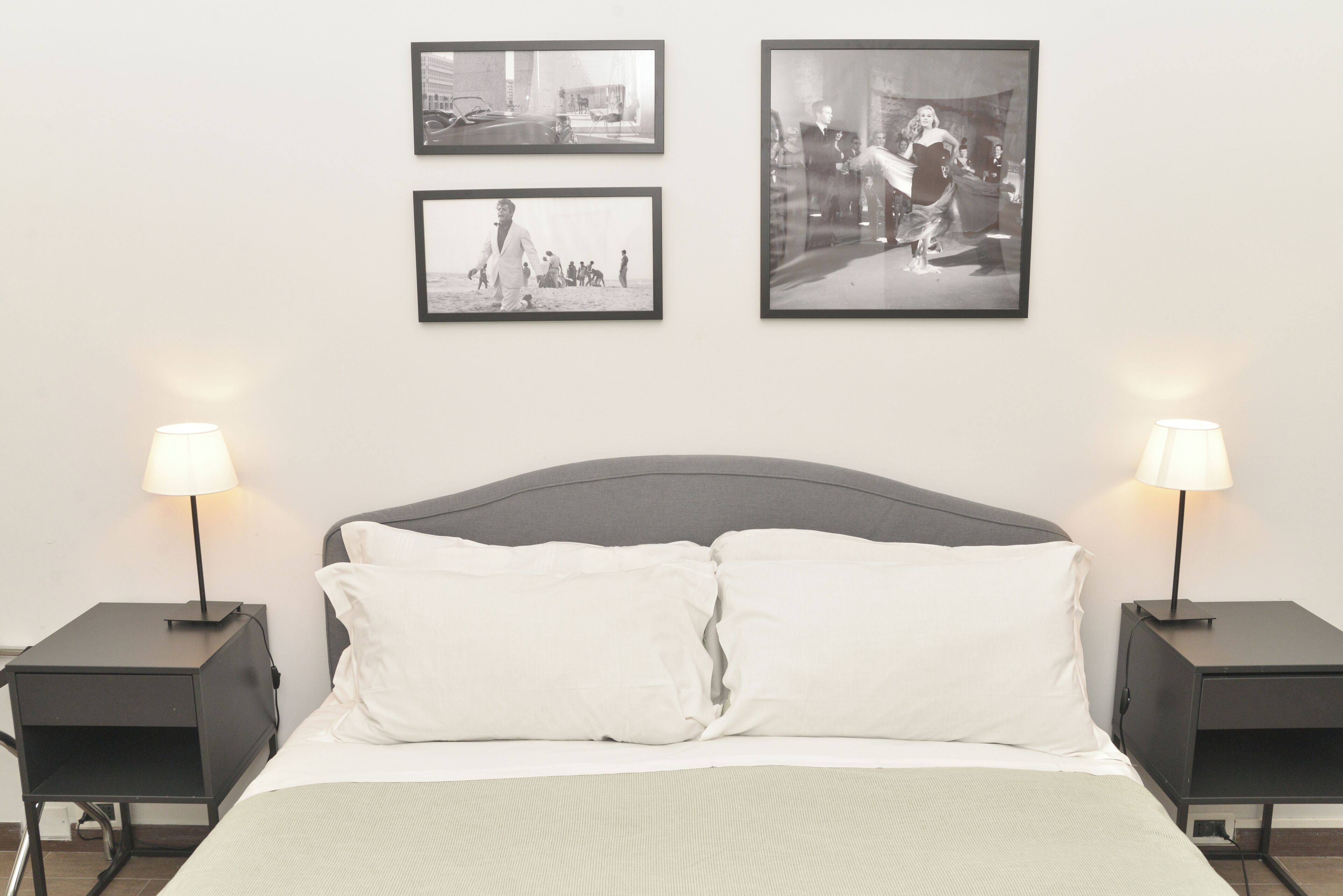 Cama bem arrumada com lençóis brancos e duas mesas de cabeceira com abajures, em quarto com três fotografias a preto e branco na parede.