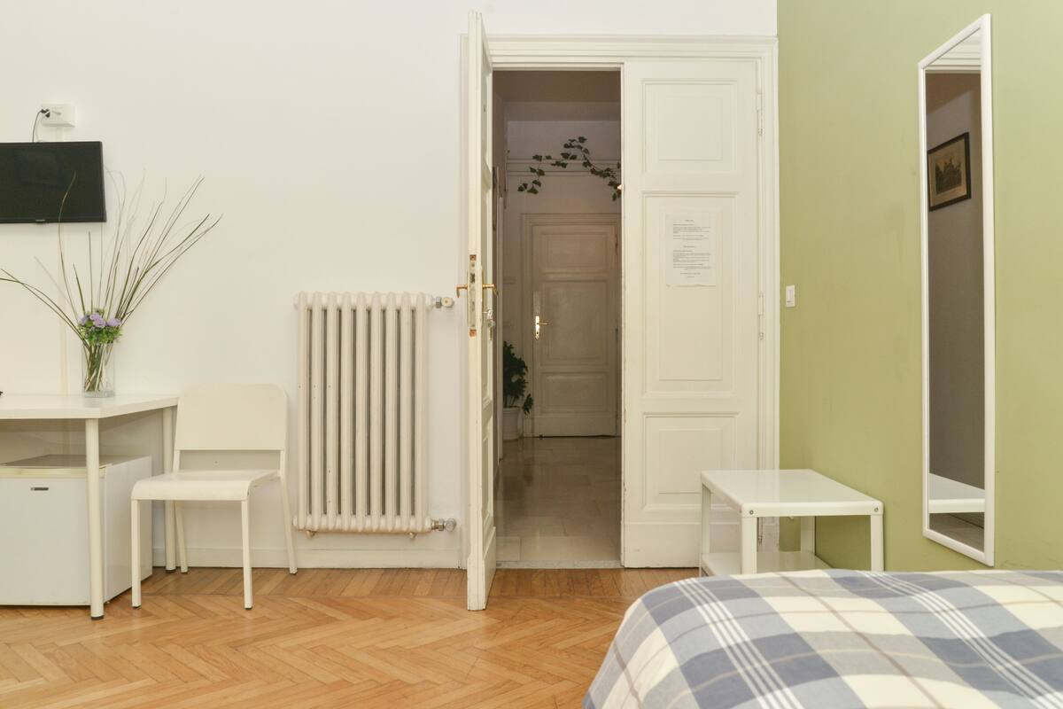 A room with a bed, a desk, a chair, a radiator, and a door leading to another room.