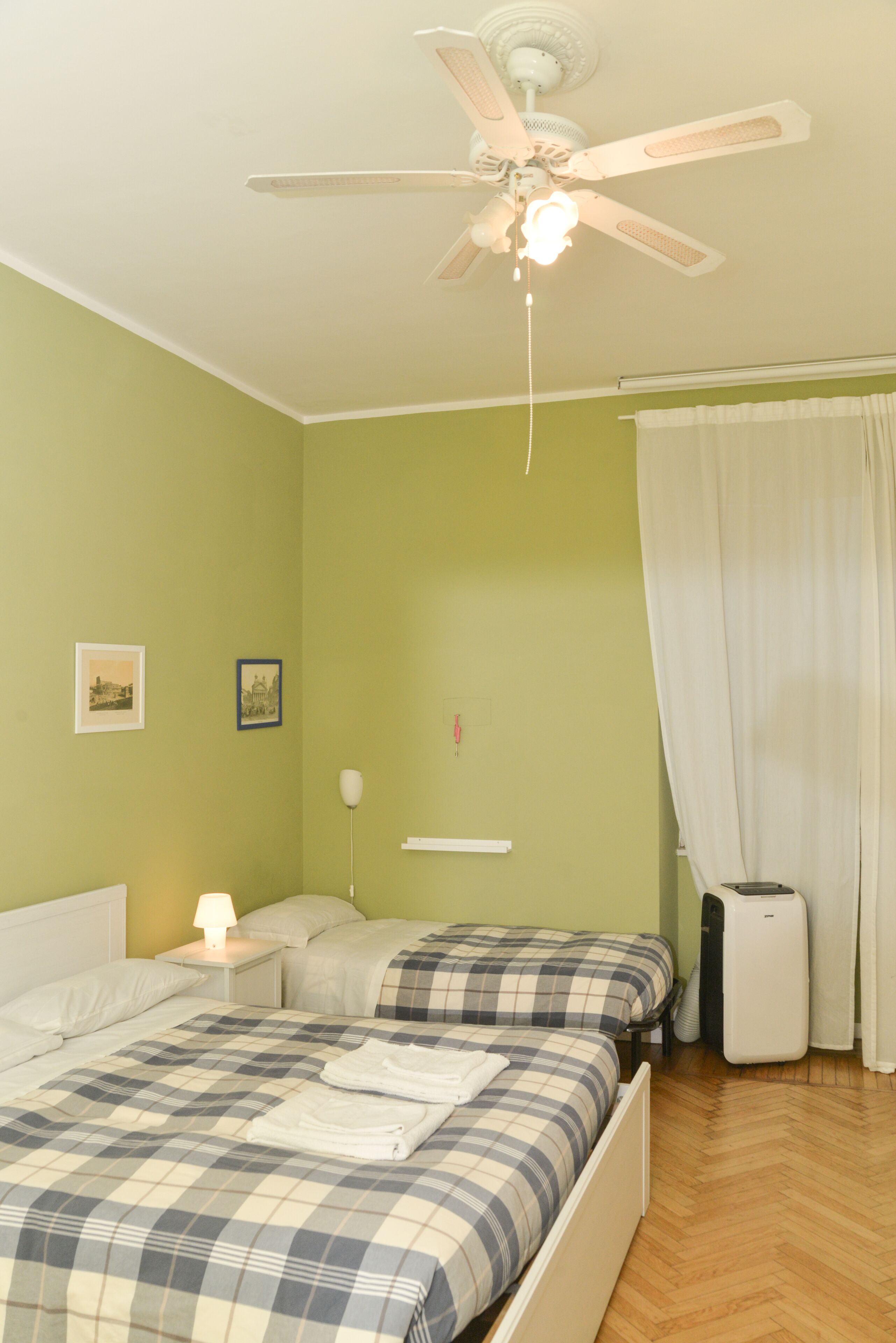 A room with two beds, a ceiling fan, a nightstand with a lamp, and a white radiator.
