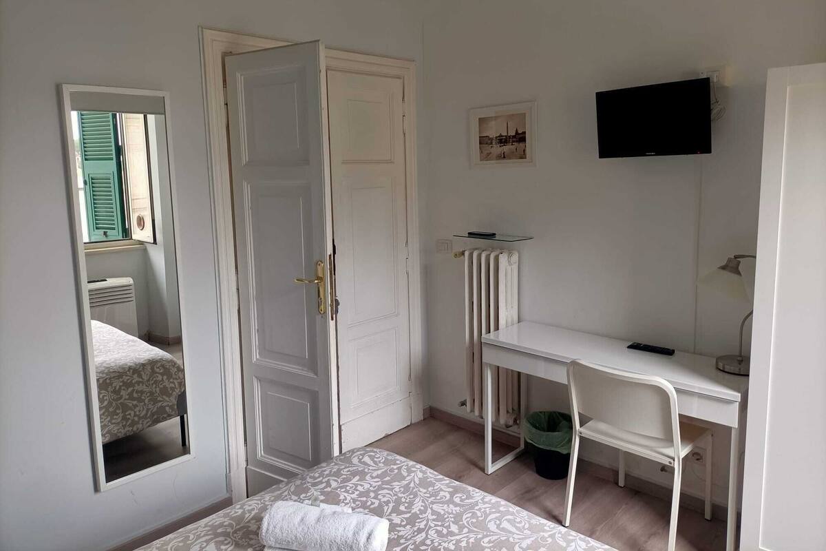 A bedroom with a bed, a desk, a chair, a radiator, a television, and a mirror.
