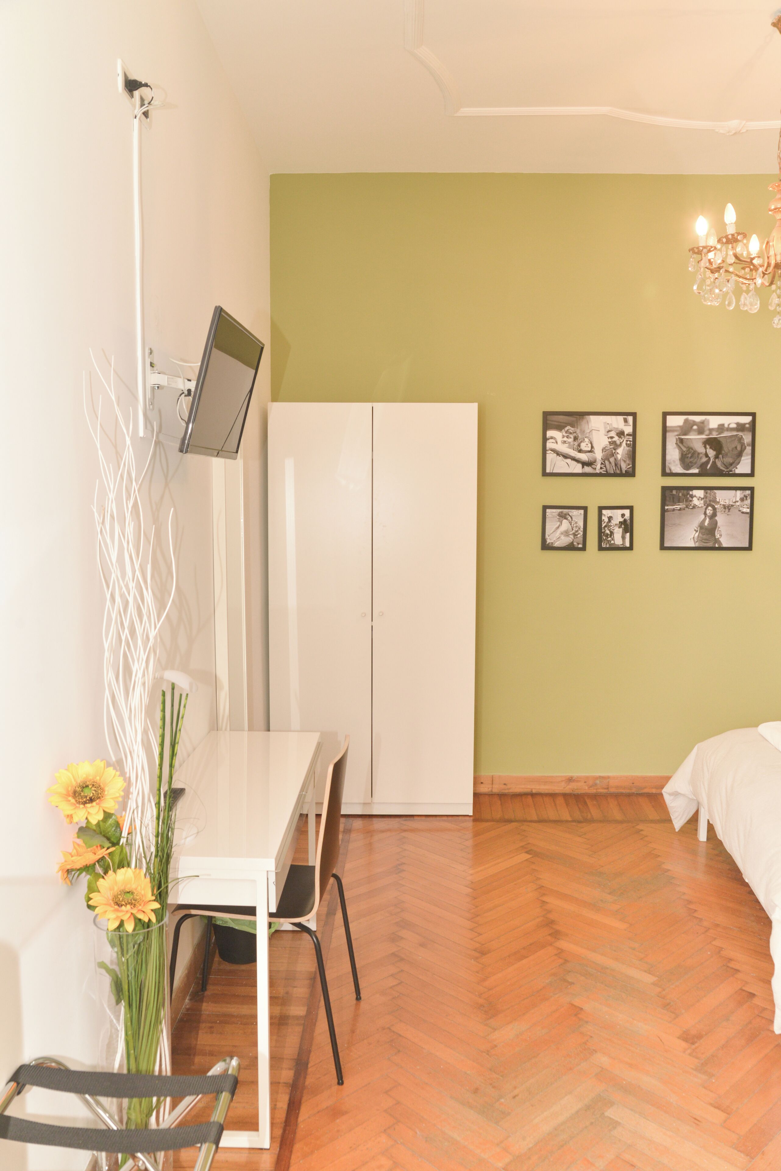A room with a wooden herringbone floor, a white desk with a chair, a white wardrobe, a bed with white bedding, a chandelier, and a wall-mounted TV.