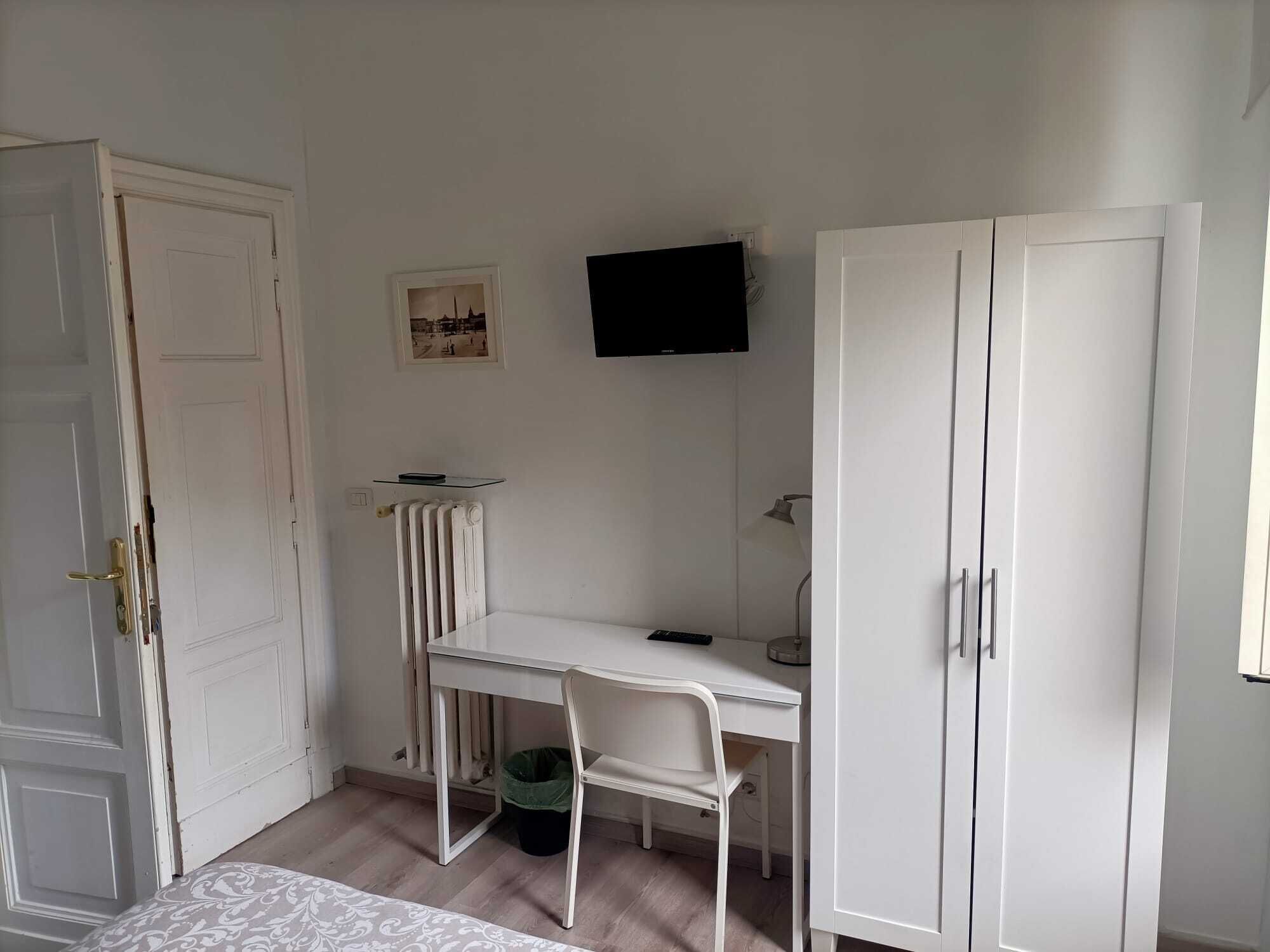 A room with a white desk, a chair, a radiator, a television, and a wardrobe.