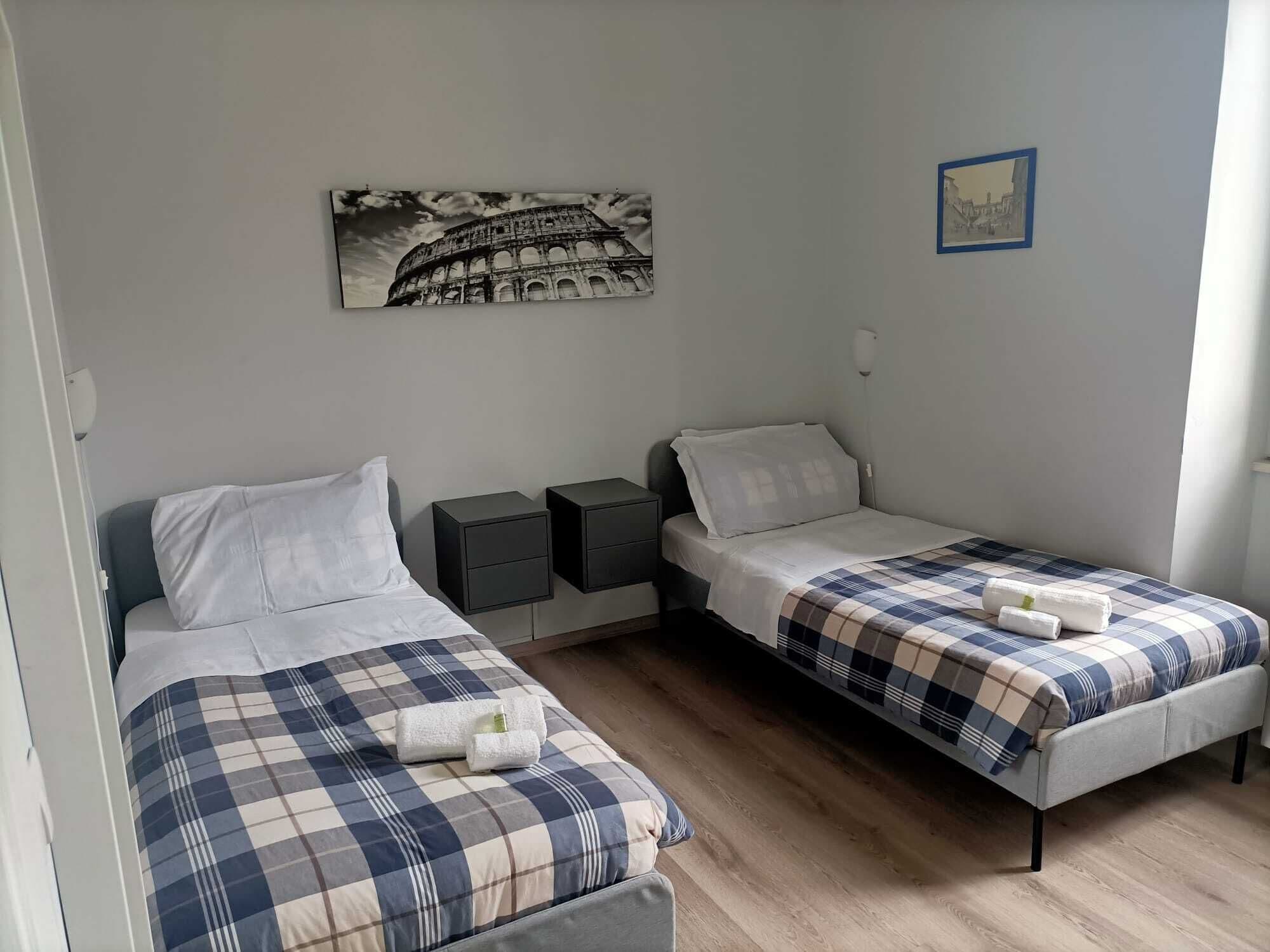 A room with two beds, each with a checkered bedspread and white pillows. There are bedside tables with drawers, a wall-mounted lamp, and a framed picture of the Colosseum.