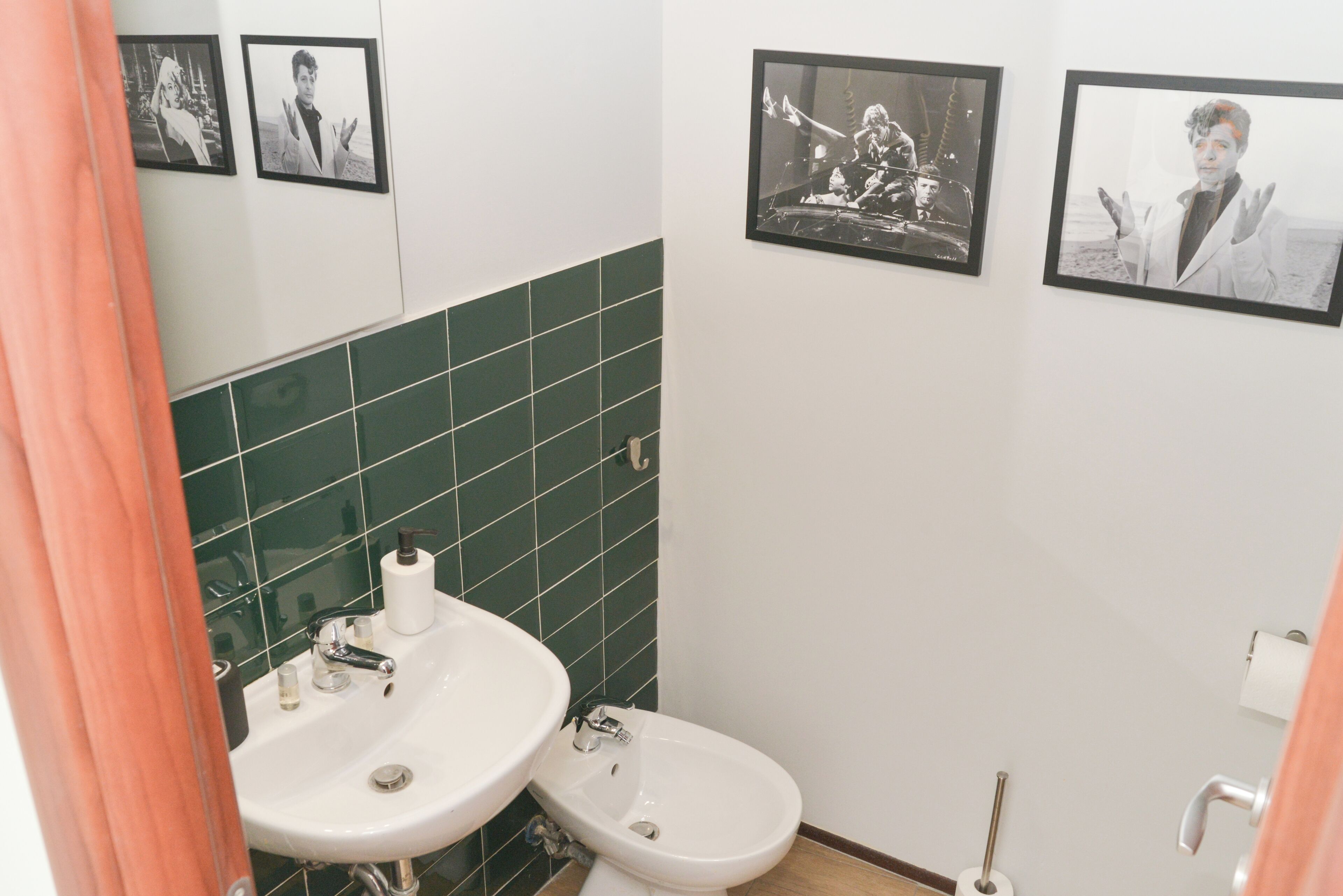 Un baño con lavamanos, inodoro y paredes de azulejos verdes. Hay cuadros enmarcados en la pared.