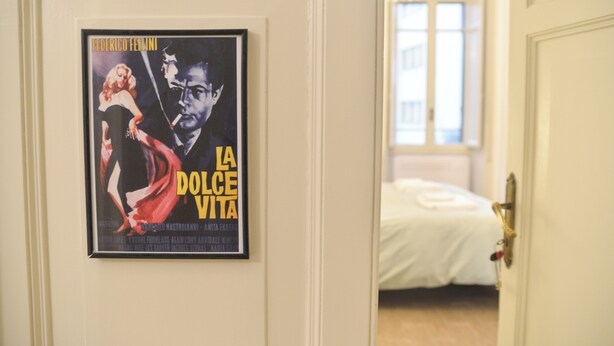 A framed movie poster on a wall, a bed with white bedding, and a door leading to another room. - Colazione Al Vaticano (Rome)