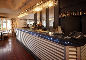 Eine Bar mit Theke, Barhocker, Kasse und Telefon. Flaschen stehen in Regalen und ein Fenster mit Vorhängen ist zu sehen.