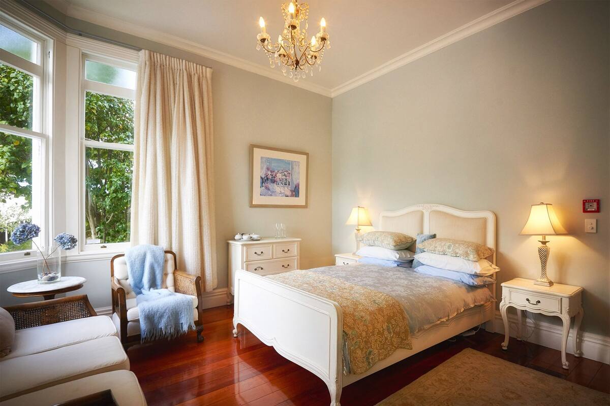 A bedroom with a large bed, two bedside tables, a chair, a small table, and a chandelier.