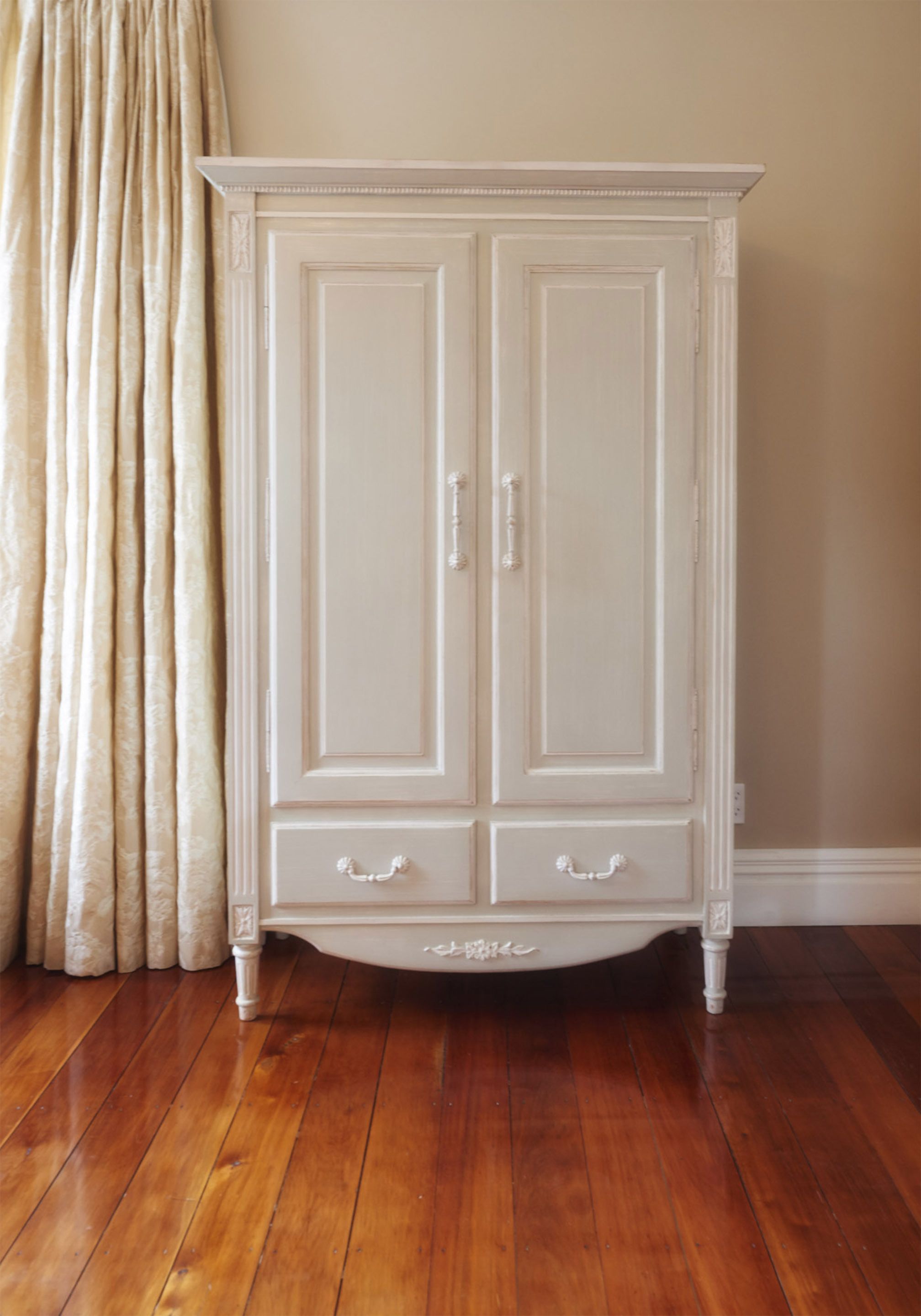 Une armoire en bois blanche, à deux portes et deux tiroirs, placée contre un mur beige et au-dessus d’un plancher en bois.