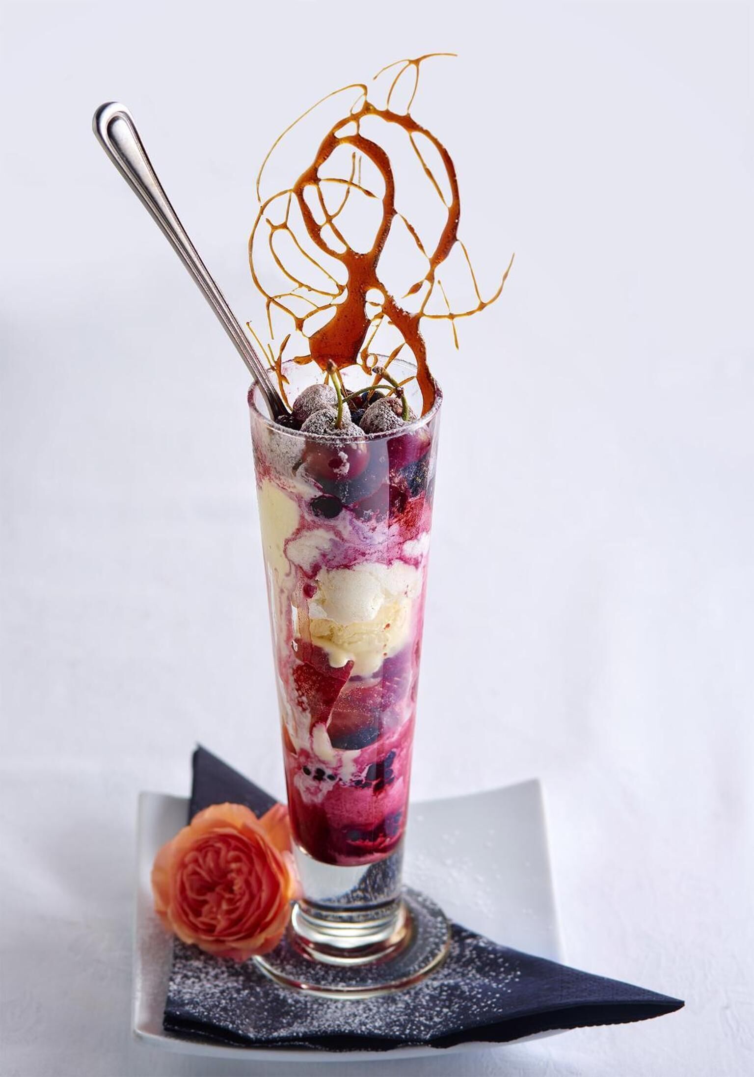 A dessert in a tall glass with a spoon, a rose, and a dark napkin.
