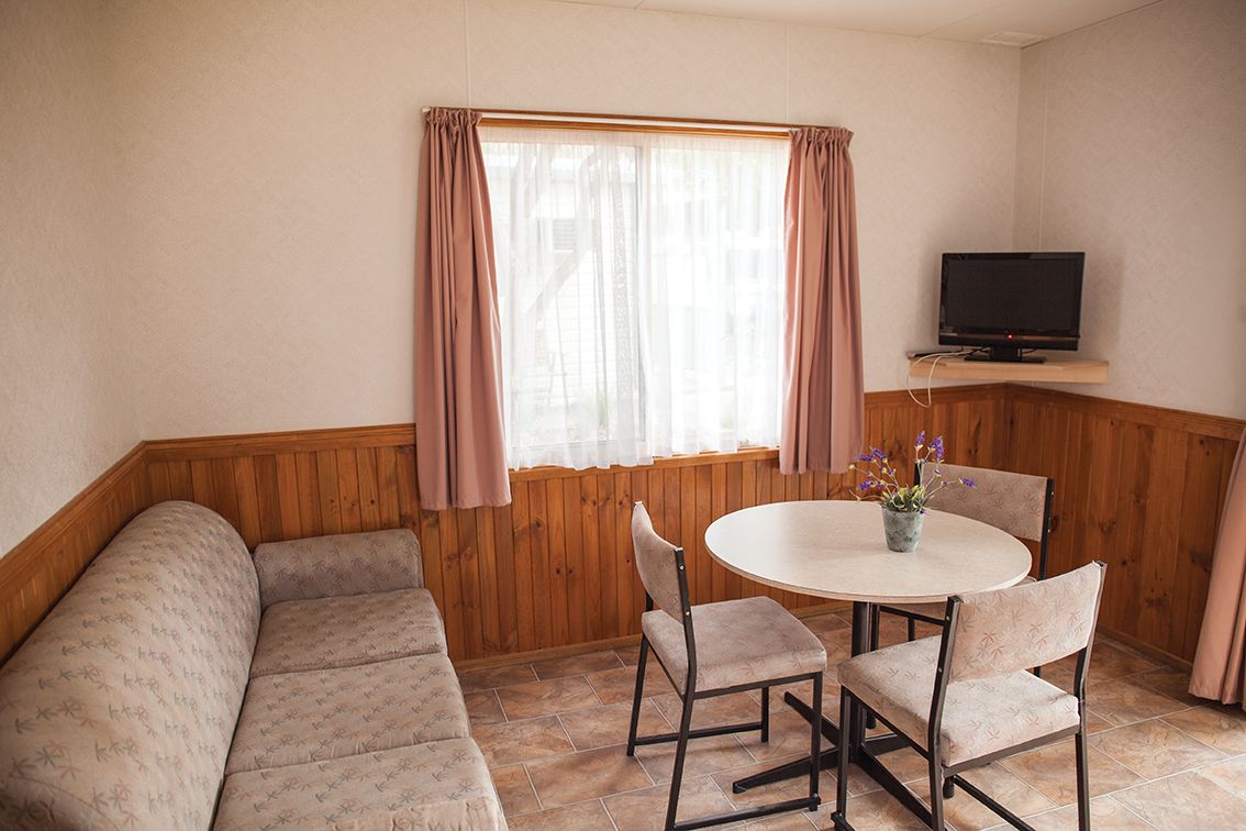 Una habitación acogedora con un banco de madera, una mesa redonda con sillas, un televisor en un estante y una ventana con cortinas.