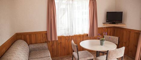 Una habitación acogedora con un banco de madera, una mesa redonda con sillas, un televisor en un estante y una ventana con cortinas.