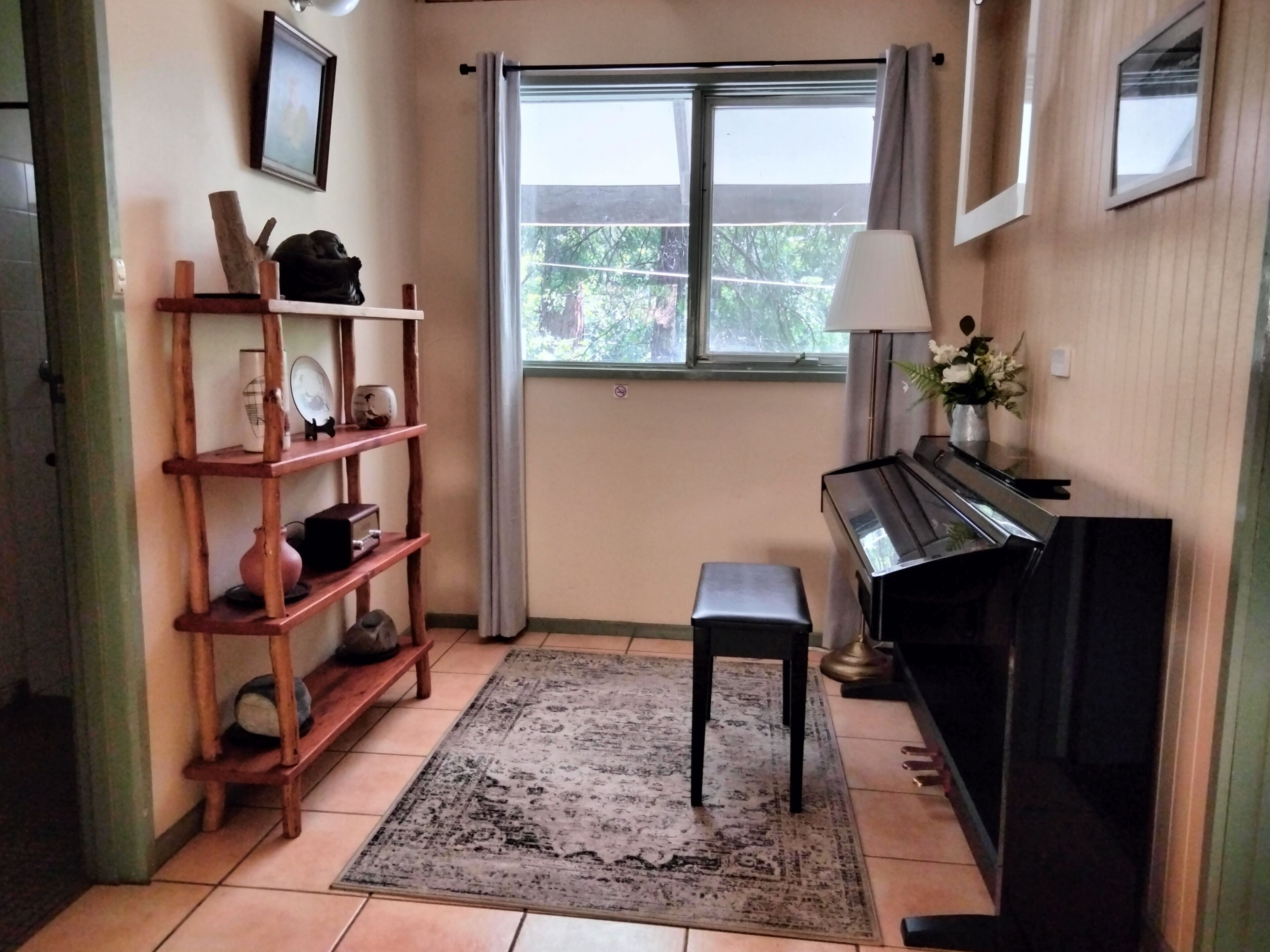 A room with a piano, a wooden shelf with decorative items, a bench, and a large window with curtains.