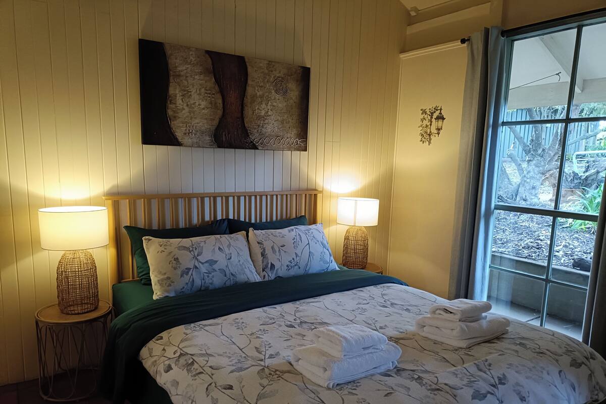 A bedroom with a bed, two bedside lamps, a window with a view of trees, and a wall-mounted artwork.