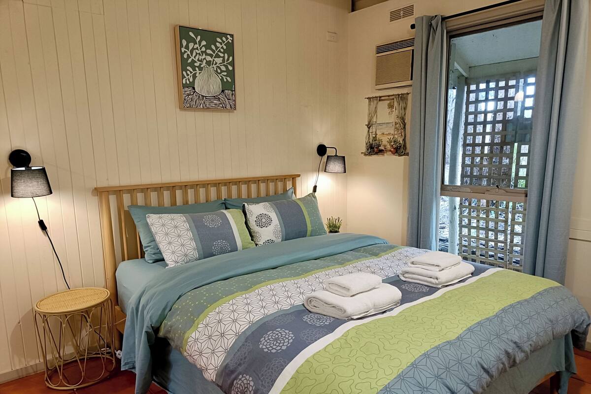A neatly made bed with patterned bedding, a bedside lamp, and a small round table with a basket.