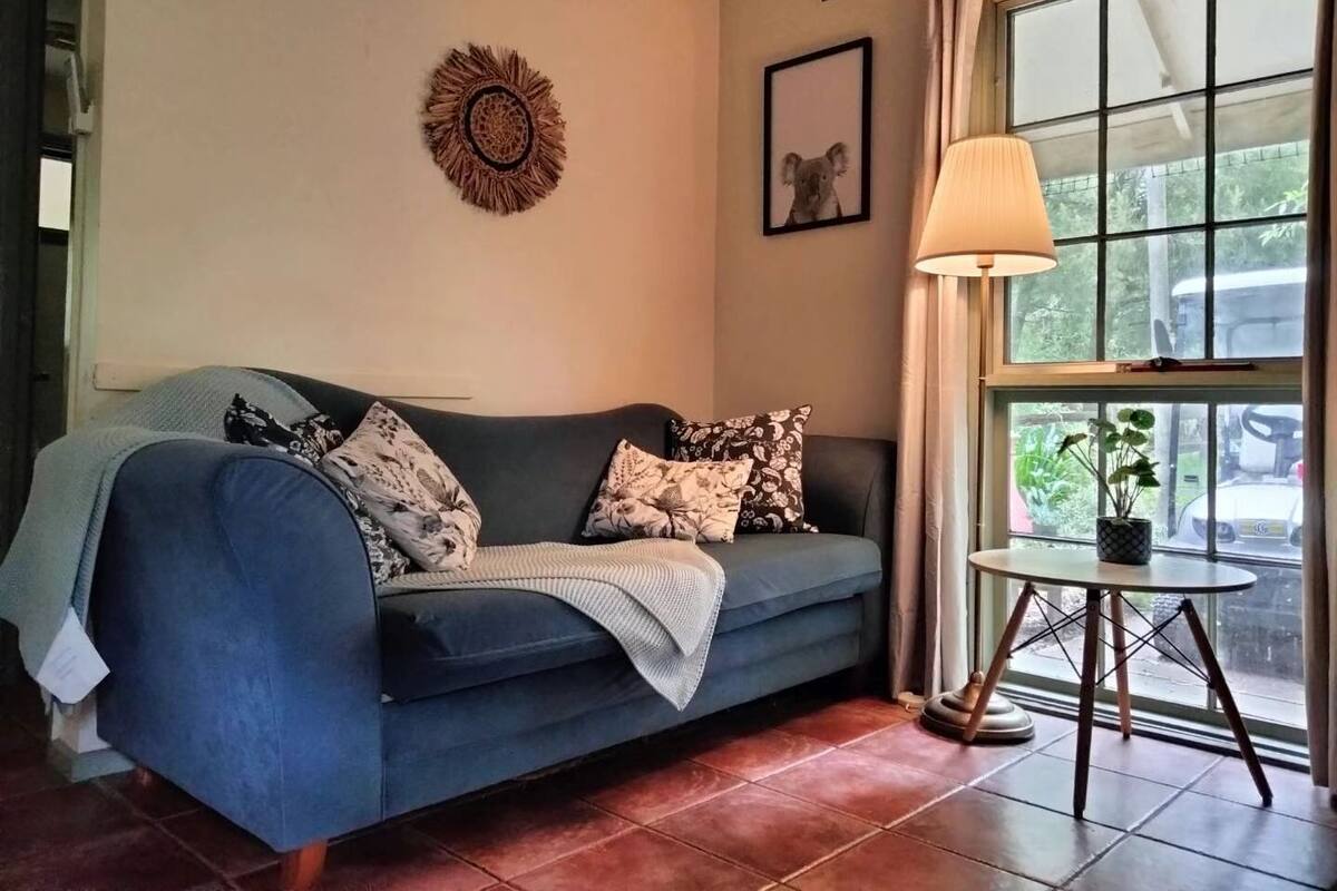 A living room with a blue sofa, a small round table with a lamp, a framed picture on the wall, and a window with curtains.