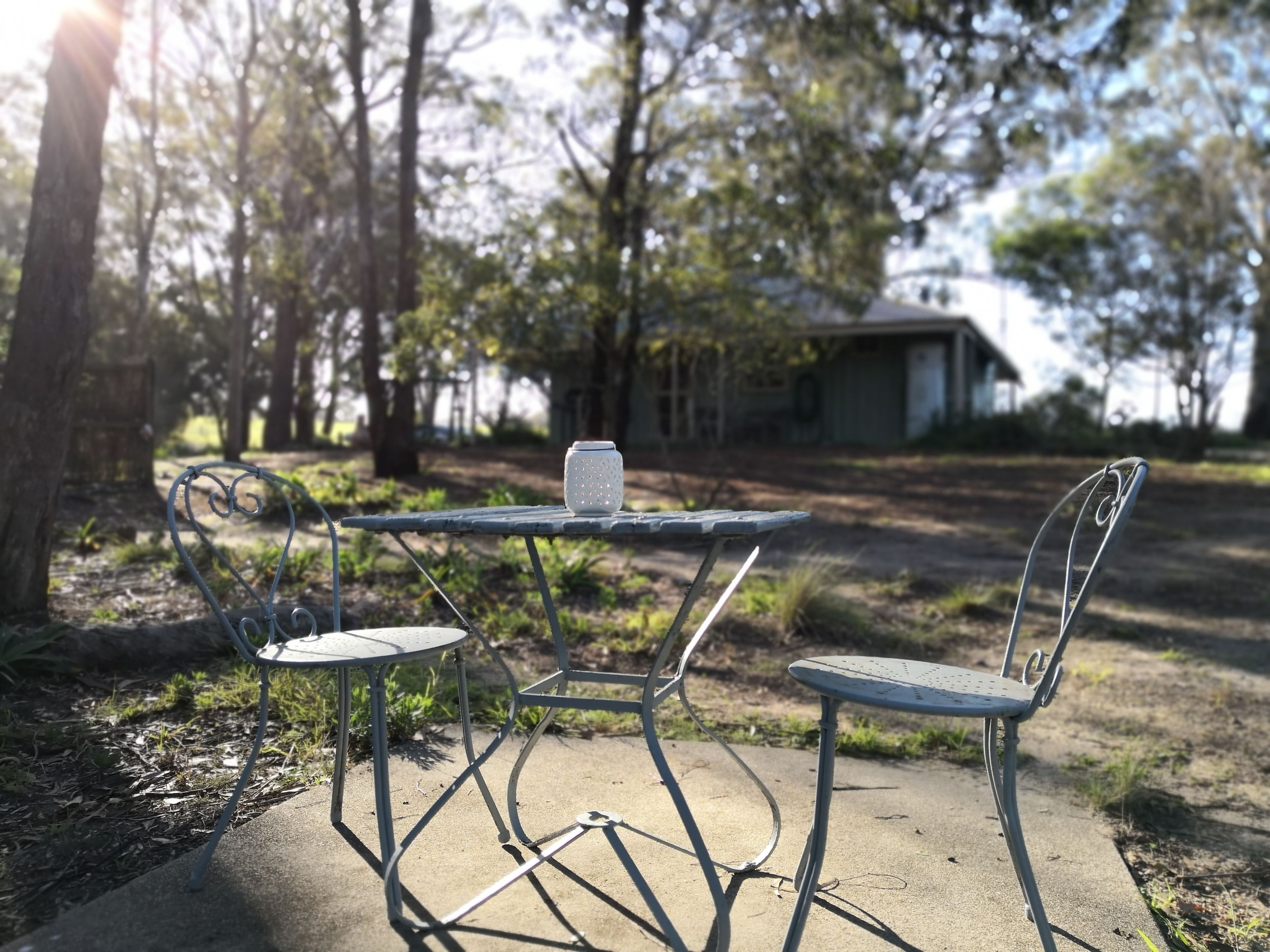 Una mesa de metal y dos sillas en un entorno exterior, con árboles y un edificio al fondo.