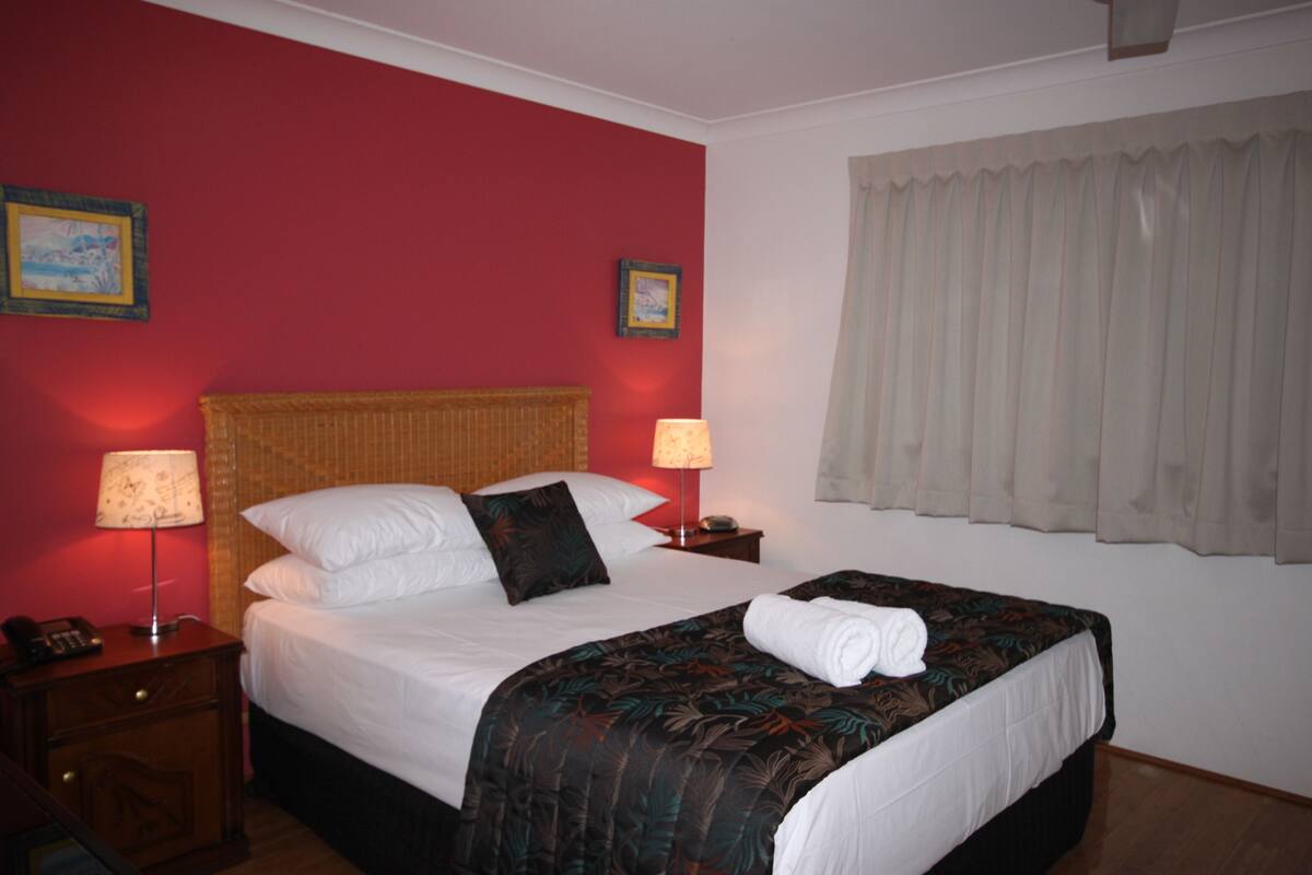 Una habitación de hotel con una cama grande, dos mesitas de noche, una pared roja y una ventana con cortinas.