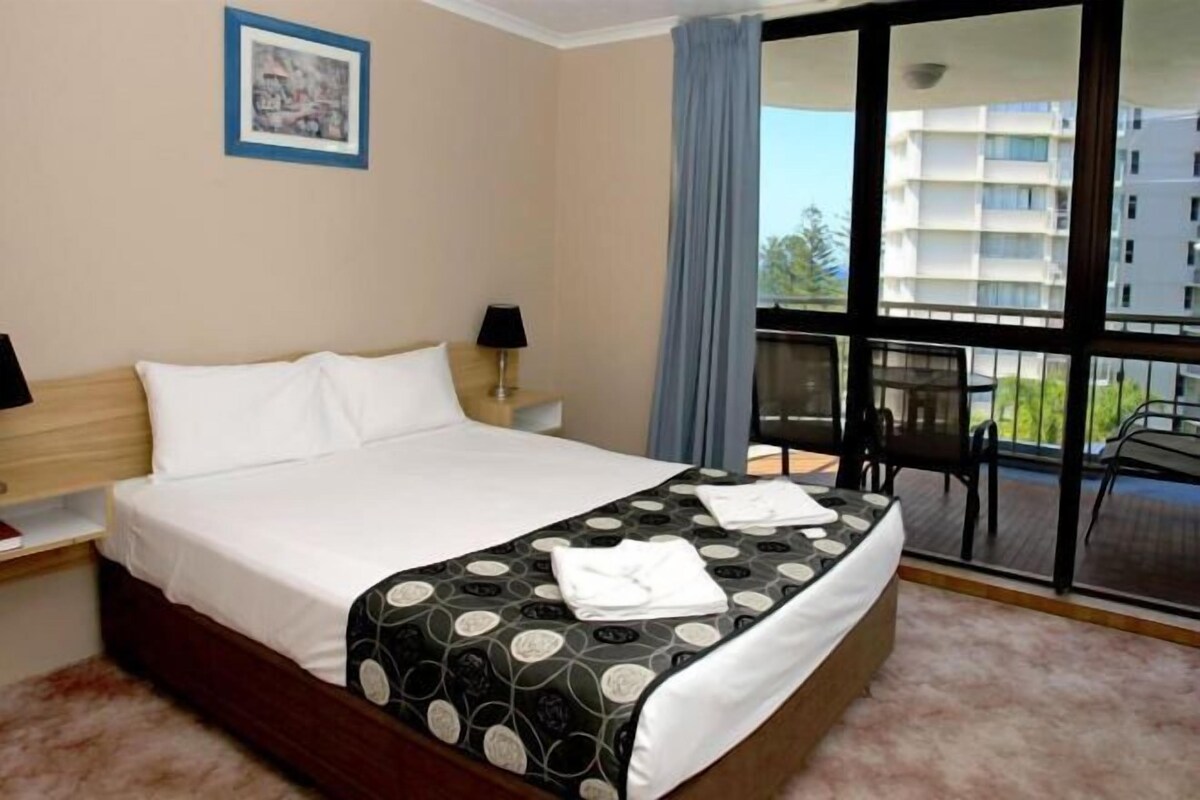 A hotel room with a bed, a nightstand, a window with a view of buildings, and a balcony with a table and chairs.