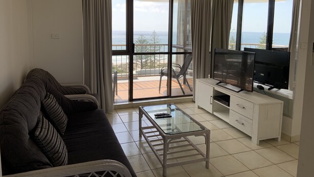 Una sala de estar con un sofá, una mesa de centro de vidrio, un televisor sobre un mueble de madera y un balcón con vista al mar.