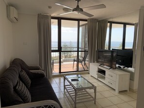 Ein Wohnzimmer mit einer Couch, einem Glastisch, einem Fernseher auf einer Holzkommode und einem Balkon mit Meerblick.