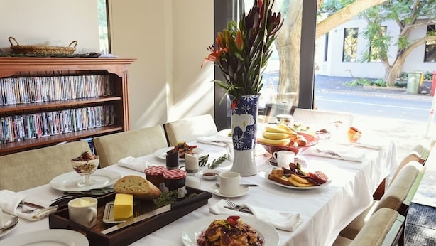 A breakfast setting with a variety of dishes, a bookshelf, and a vase of flowers. - Lily Sands Inn (South Melbourne)